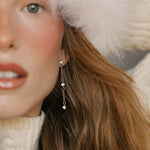 A close-up of a woman with wavy brown hair wearing a white fuzzy hat, white knit sweater, and LLUME Jewelry’s Reverie Earrings—elegant drops that shimmer. She has light skin and natural makeup with rosy cheeks.