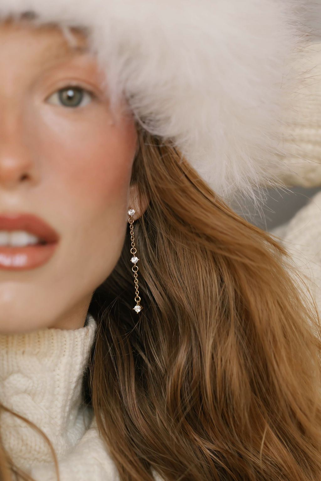 A close-up of a woman with wavy brown hair wearing a white fuzzy hat, white knit sweater, and LLUME Jewelry’s Reverie Earrings—elegant drops that shimmer. She has light skin and natural makeup with rosy cheeks.