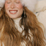 A joyful woman with long wavy hair and a bright smile wears Reverie Earrings by LLUME Jewelry, a fluffy white hat, and cream knit sweater, eyes closed and arms raised behind her head, radiating warmth and happiness.