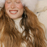 A joyful woman with long wavy hair and a bright smile wears Reverie Earrings by LLUME Jewelry, a fluffy white hat, and cream knit sweater, eyes closed and arms raised behind her head, radiating warmth and happiness.