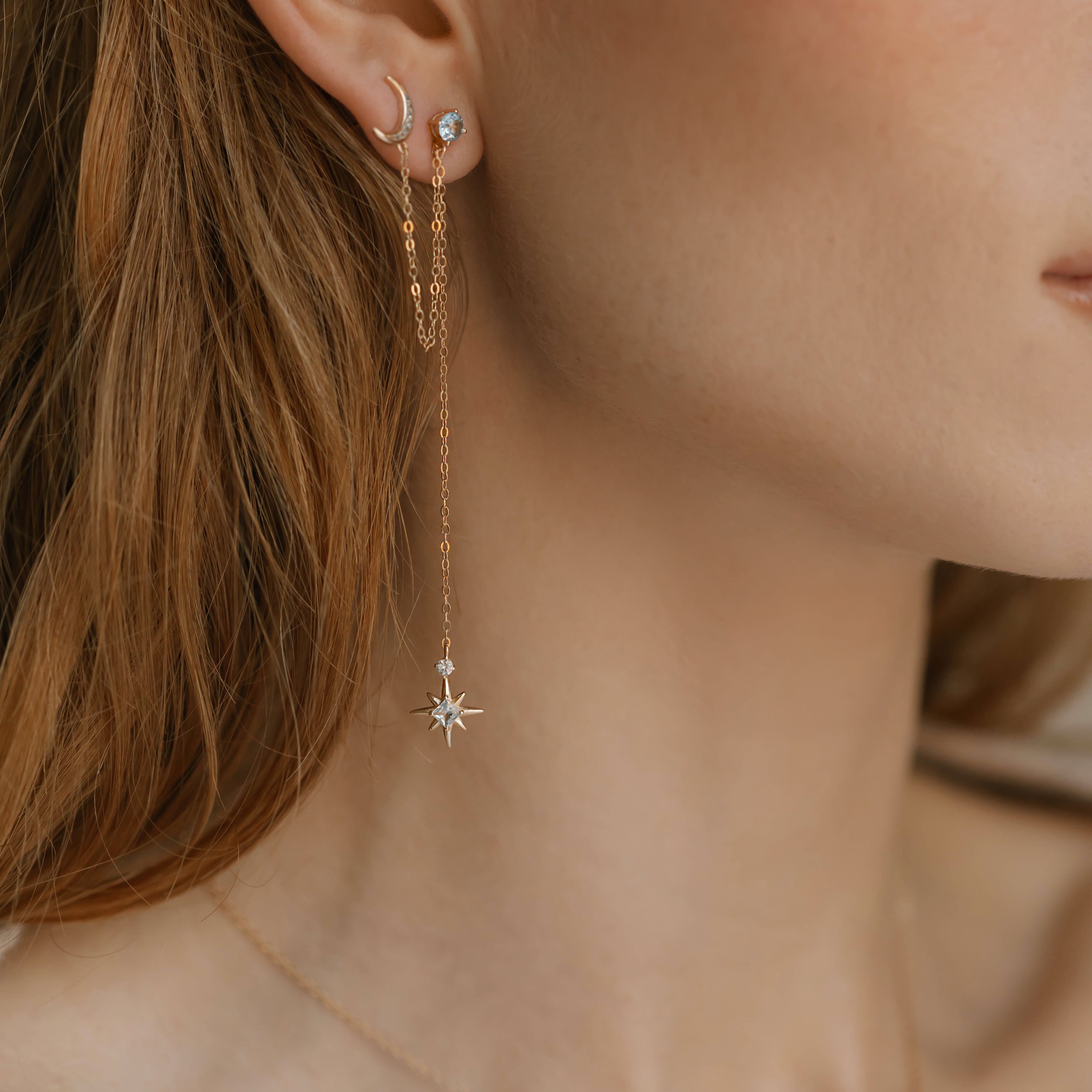 A close-up of a woman’s ear and neck features LLUME Jewelry's Night Sky Earrings—beautiful gold pieces with crescent moon and star details—layered elegantly, ideal for winter style.