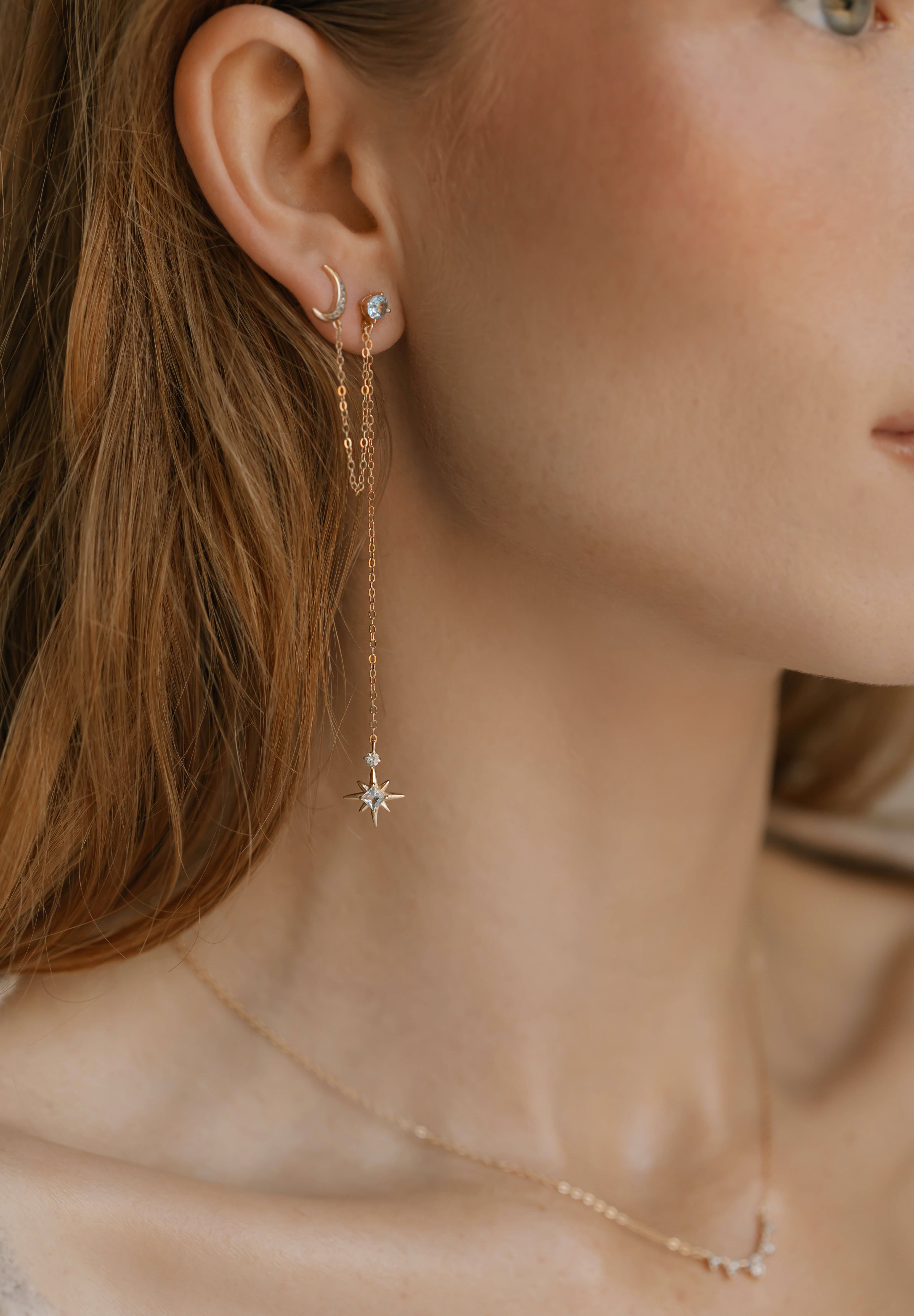 A close-up of a woman’s ear and neck features LLUME Jewelry's Night Sky Earrings—beautiful gold pieces with crescent moon and star details—layered elegantly, ideal for winter style.