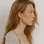 A young woman with long, wavy, light brown hair wears a light gray sweater, delicate jewelry, and LLUME Jewelry's Night Sky Earrings—a perfect choice for elegant winter style.