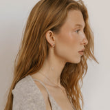 A young woman with long, wavy, light brown hair wears a light gray sweater, delicate jewelry, and LLUME Jewelry's Night Sky Earrings—a perfect choice for elegant winter style.