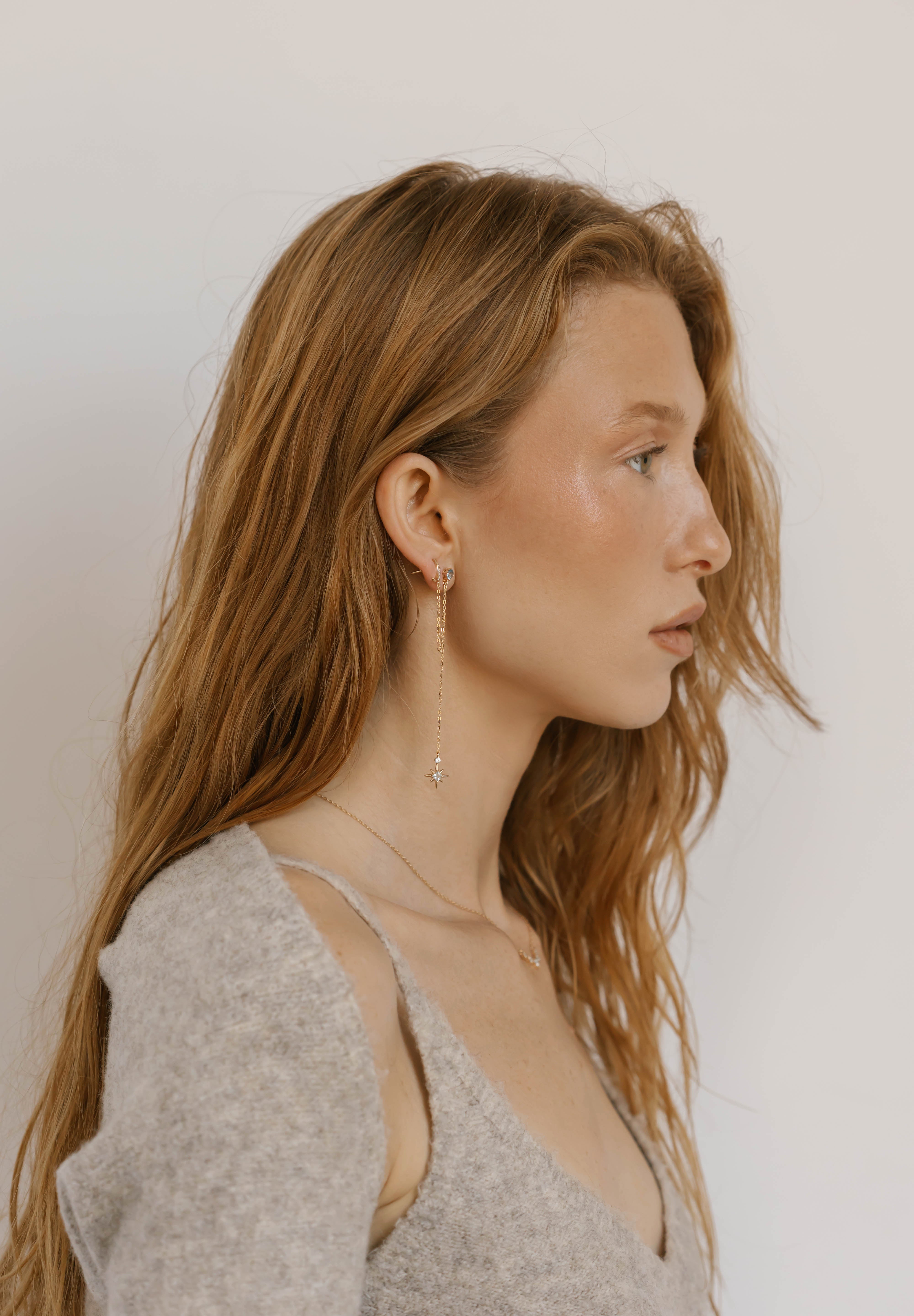 A young woman with long, wavy, light brown hair wears a light gray sweater, delicate jewelry, and LLUME Jewelry's Night Sky Earrings—a perfect choice for elegant winter style.