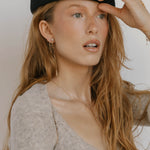 A woman with long, wavy red hair wears a black beret, a beige fuzzy top, and LLUME Jewelry's Night Sky Earrings. She gazes to the side with her mouth slightly open, holding her hat—an effortlessly chic winter look.