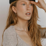 A woman with long, wavy red hair wears a black beret, a beige fuzzy top, and LLUME Jewelry's Night Sky Earrings. She gazes to the side with her mouth slightly open, holding her hat—an effortlessly chic winter look.
