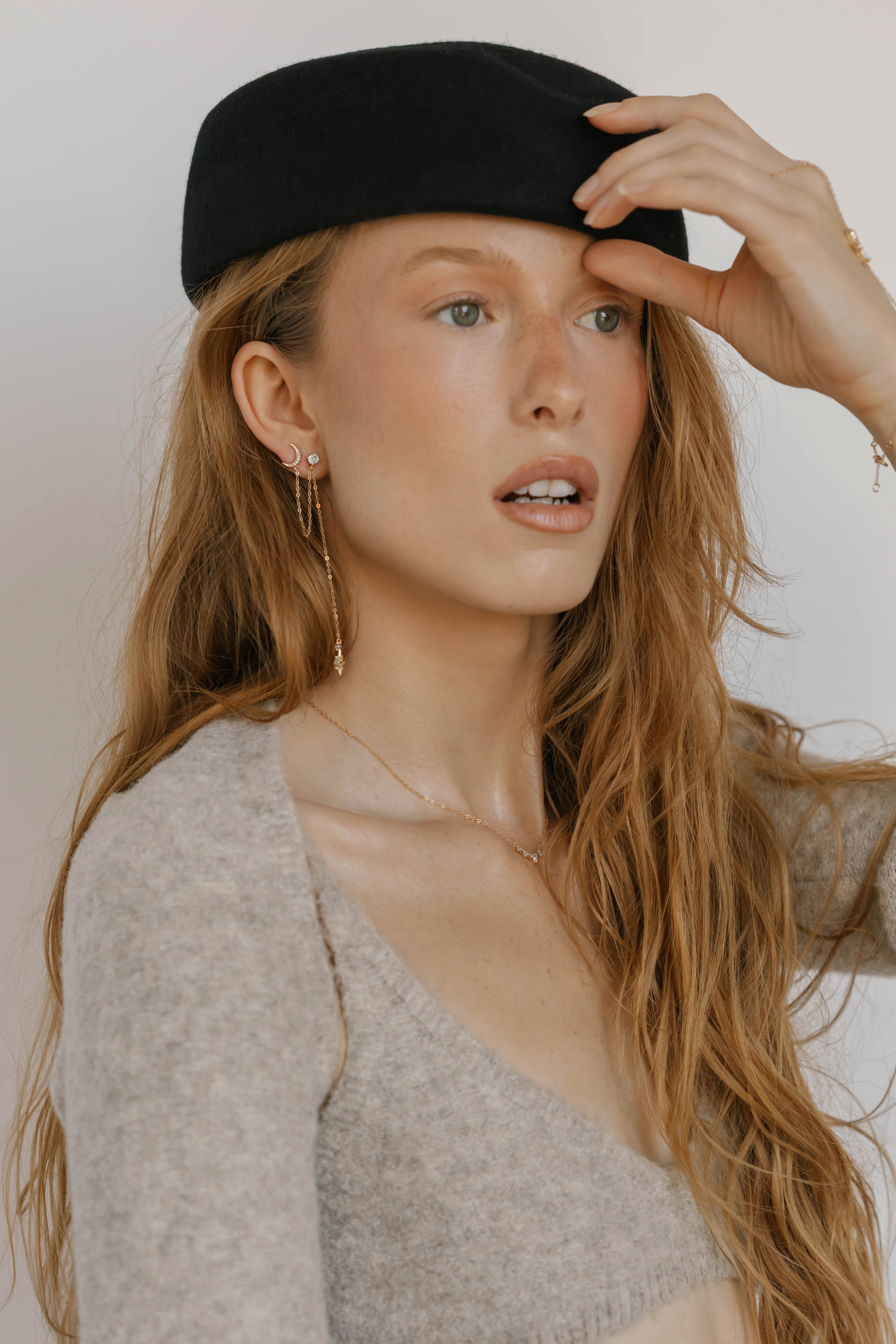A woman with long, wavy red hair wears a black beret, a beige fuzzy top, and LLUME Jewelry's Night Sky Earrings. She gazes to the side with her mouth slightly open, holding her hat—an effortlessly chic winter look.