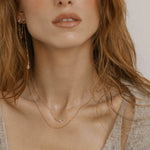 A woman with wavy, light brown hair wears dainty gold necklaces and LLUME Jewelry's Night Sky Earrings. In a soft beige sweater, she effortlessly captures winter style from shoulders to lips.