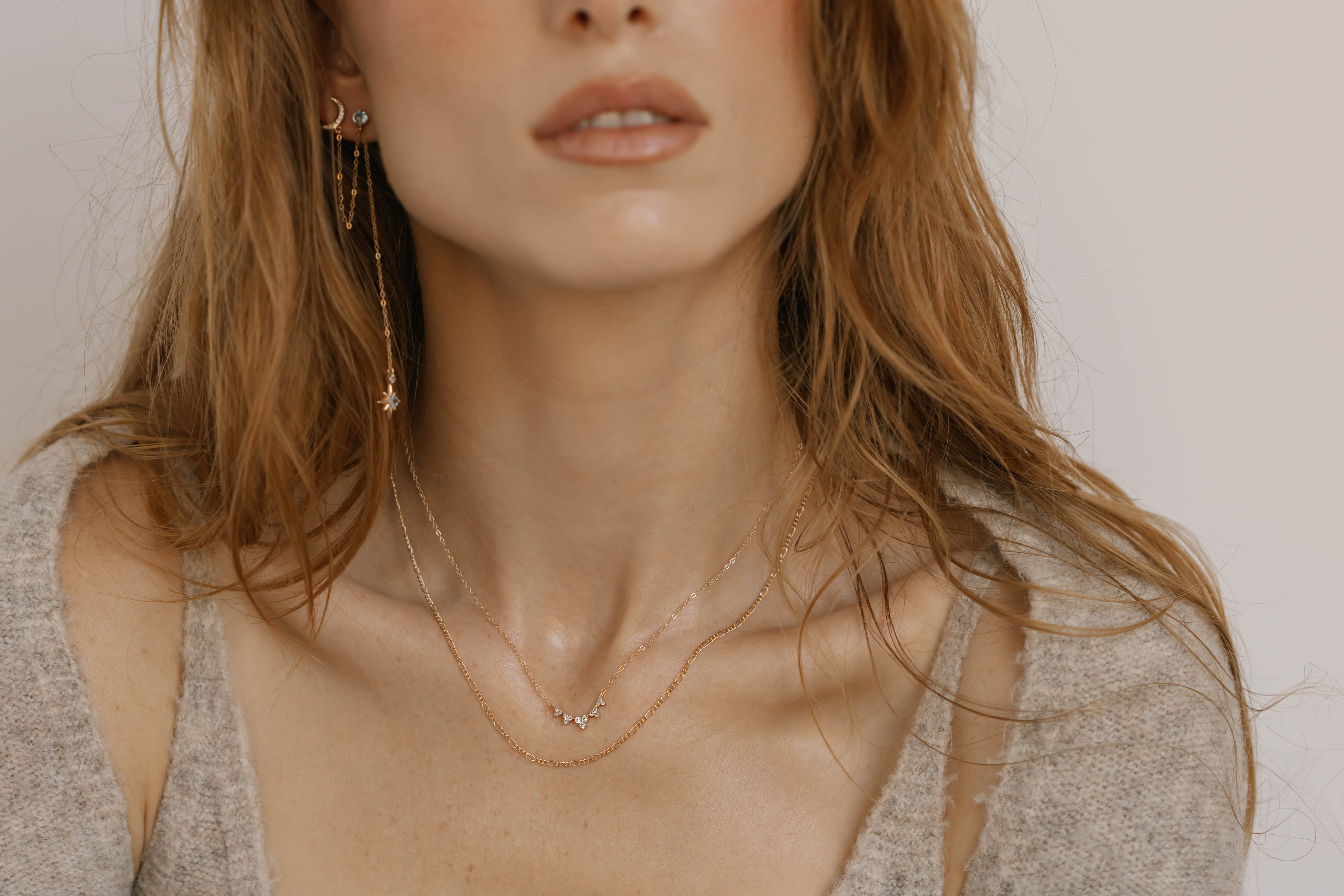 A woman with wavy, light brown hair wears dainty gold necklaces and LLUME Jewelry's Night Sky Earrings. In a soft beige sweater, she effortlessly captures winter style from shoulders to lips.