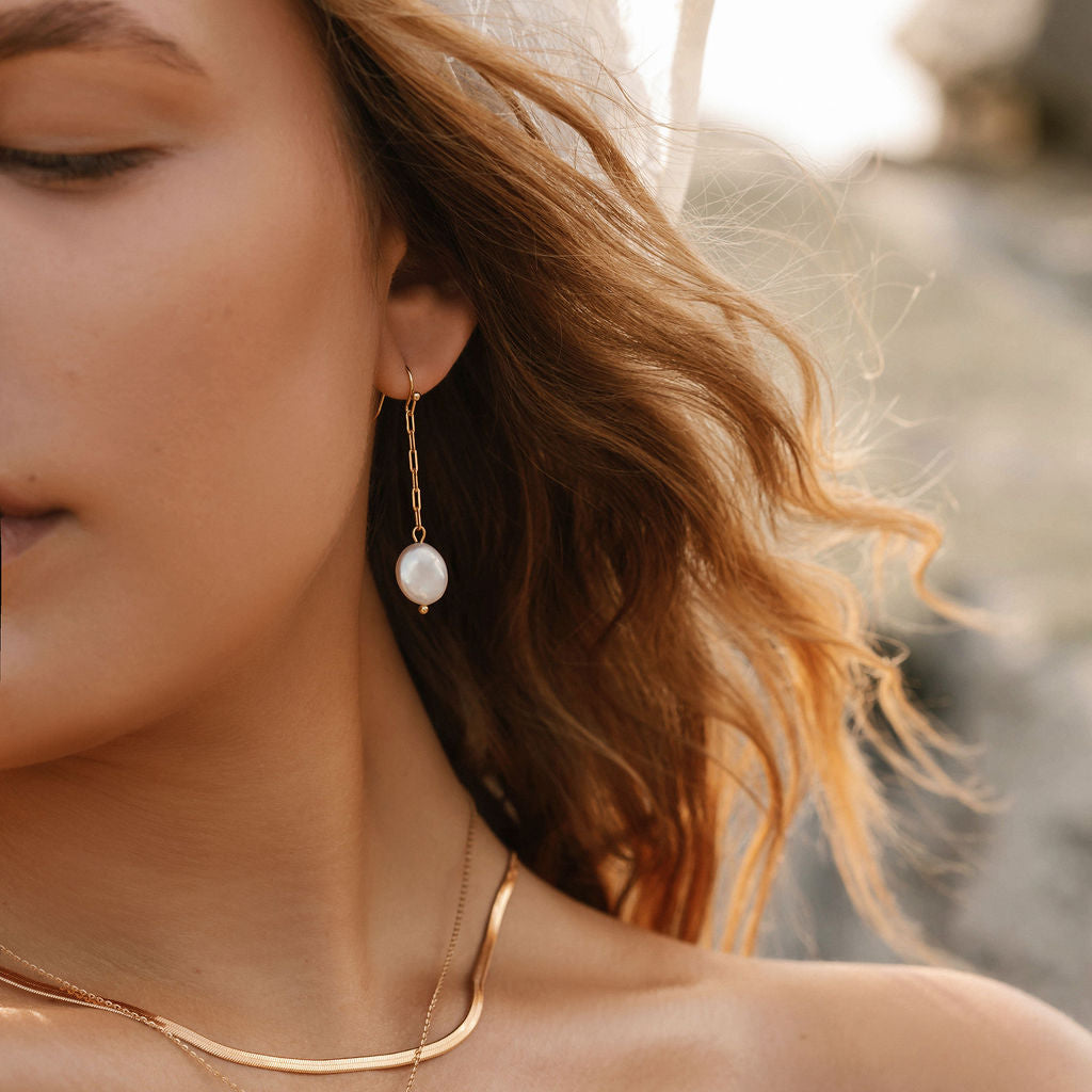 Close-up of a woman with wavy hair wearing the Lara Pearl Earrings by LLUME Jewelry, along with gold chain necklaces and a sheer, embroidered veil. Sunlight casts a warm glow, with softly blurred rocks in the background.