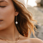 Close-up of a woman with wavy hair wearing the Lara Pearl Earrings by LLUME Jewelry, along with gold chain necklaces and a sheer, embroidered veil. Sunlight casts a warm glow, with softly blurred rocks in the background.