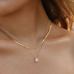 A close-up of a woman’s neck wearing The Classic Set by LLUME Jewelry, featuring a gold herringbone chain, a pendant necklace with a square charm, and a single dangling pearl earring.