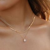 A close-up of a woman’s neck wearing The Classic Set by LLUME Jewelry, featuring a gold herringbone chain, a pendant necklace with a square charm, and a single dangling pearl earring.