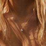 A close-up of a woman wearing LLUME Jewelry’s The One Necklace—a gold plated piece with a square pendant and clear gem charm—against her bare skin, wavy blonde hair, and a textured white top.