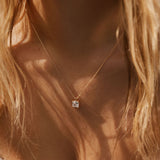 A close-up of a woman wearing LLUME Jewelry’s The One Necklace—a gold plated piece with a square pendant and clear gem charm—against her bare skin, wavy blonde hair, and a textured white top.