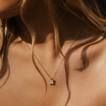 Close-up of a woman's neck and collarbone, with wavy hair, wearing LLUME Jewelry's The One Necklace—a gold plated piece with a square-cut clear gem pendant—styled with a white, off-the-shoulder top.