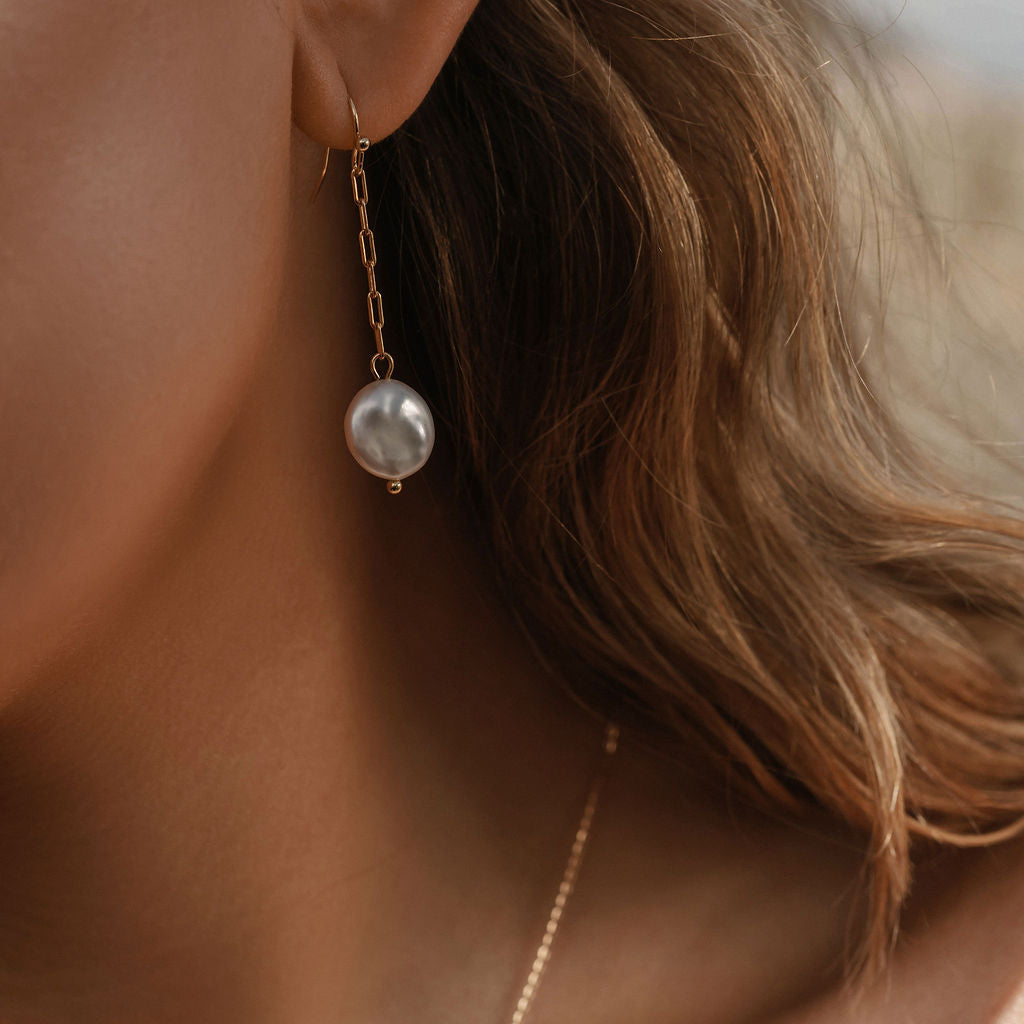 Close-up of a woman with light brown hair wearing LLUME Jewelry’s Lara Pearl Earrings—elegant gold-plated pearls. Part of a gold necklace appears on her neck, and the softly blurred background highlights the refined accessory.