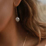 Close-up of a woman with light brown hair wearing LLUME Jewelry’s Lara Pearl Earrings—elegant gold-plated pearls. Part of a gold necklace appears on her neck, and the softly blurred background highlights the refined accessory.