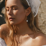 A young woman with closed eyes and long blonde hair wears LLUME Jewelry's Lara Pearl Earrings, a white lace headscarf, a delicate necklace, and an off-the-shoulder white top as she stands outdoors in the sunlight.