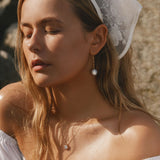 A young woman with closed eyes and long blonde hair wears LLUME Jewelry's Lara Pearl Earrings, a white lace headscarf, a delicate necklace, and an off-the-shoulder white top as she stands outdoors in the sunlight.