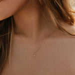 A close-up of a woman's neck wearing the LLUME Jewelry Whisper Necklace—a gold-plated sterling silver piece with a small rectangular pendant—against her light skin, white off-shoulder top, and partially visible loose hair.