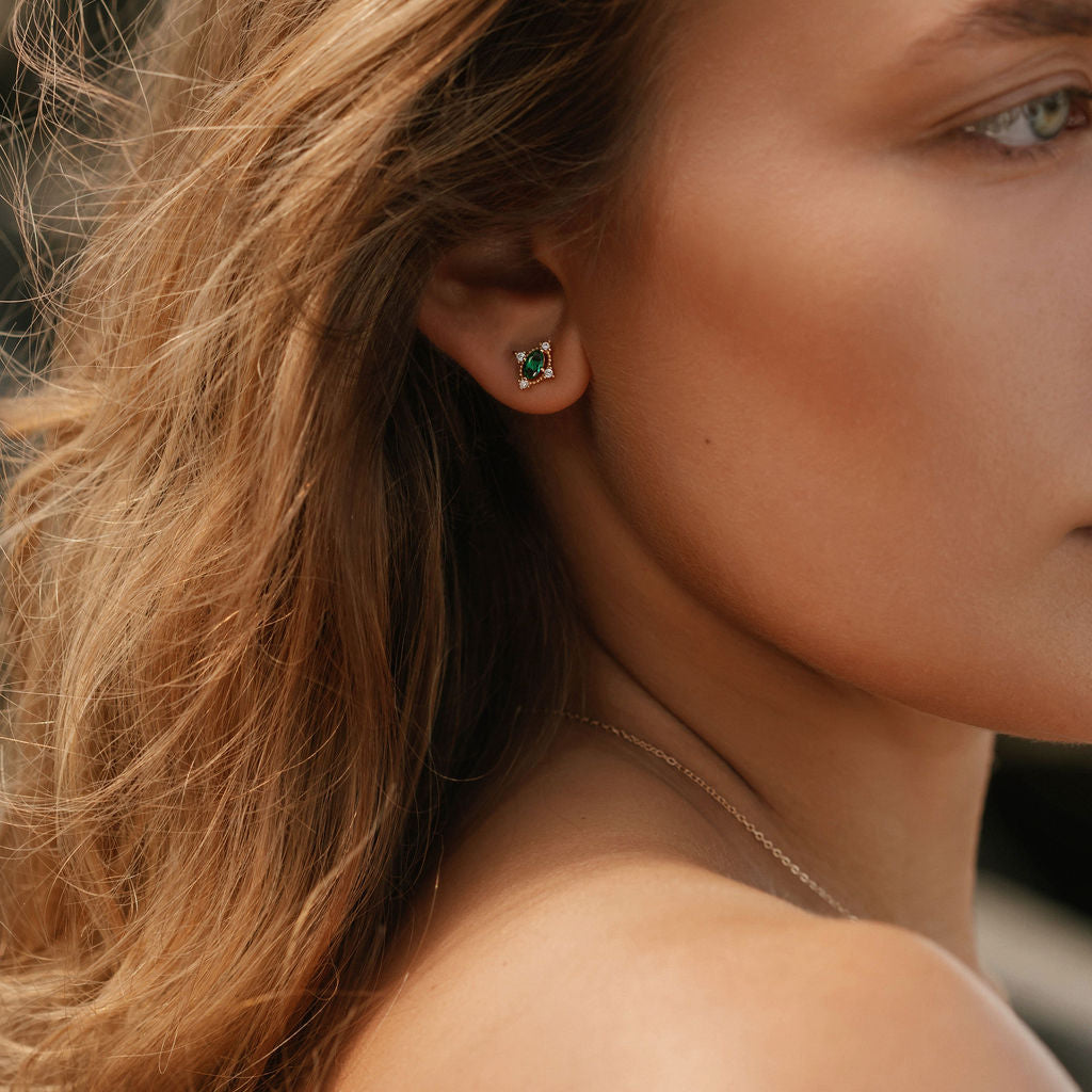 Close-up of a woman with wavy light brown hair wearing LLUME Jewelry’s Destiny Earrings—emerald green studs—and a thin necklace, set against a blurred background.