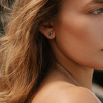 Close-up of a woman with wavy light brown hair wearing LLUME Jewelry’s Destiny Earrings—emerald green studs—and a thin necklace, set against a blurred background.