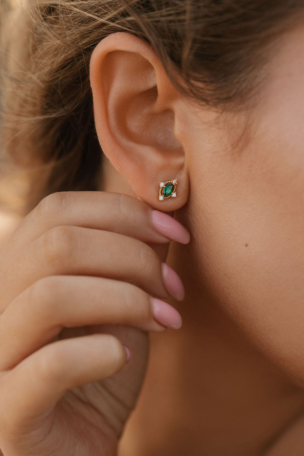 A close-up shows a person wearing LLUME Jewelry’s Destiny Earrings—small gold studs with emerald green gemstones encircled by clear stones—while their pink-manicured hand gently touches their ear.