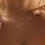 A close-up of a woman wearing the LLUME Jewelry Whisper Daily Set—two delicate gold necklaces, one with a subtle pendant—against bare skin and a white strapless top. Her hands gently touch the chains, highlighting timeless everyday style.