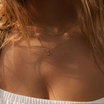 Close-up of a woman with long blonde hair in an off-shoulder white top wearing the LLUME Jewelry Whisper Daily Set—two delicate gold necklaces, including a small vertical Whisper pendant—perfect for everyday wear.