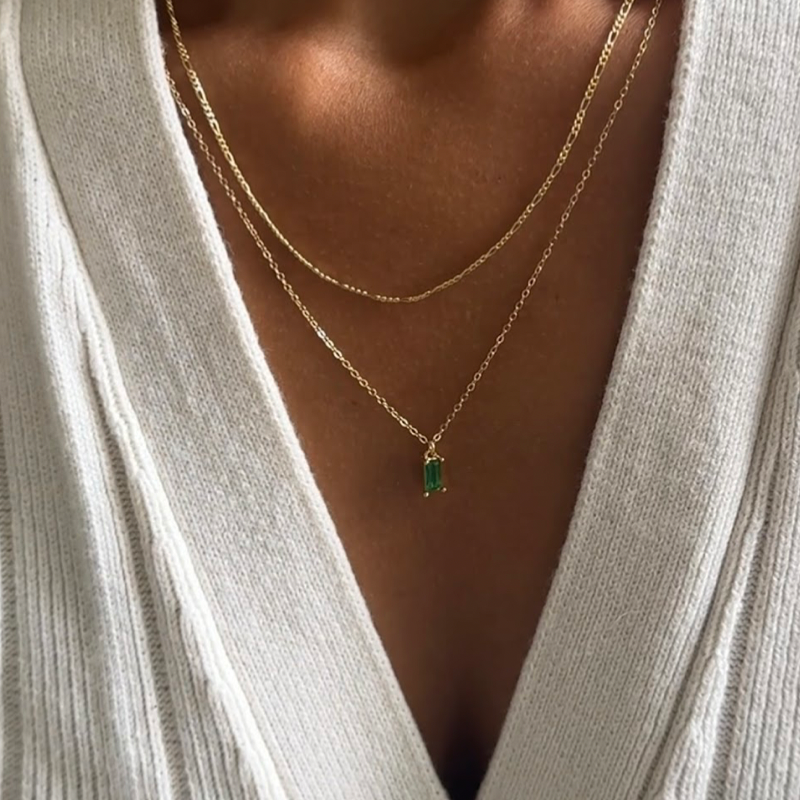A person wearing a cream ribbed sweater with a deep V-neck, showing two gold necklaces. The shorter chain is plain, and the longer chain features a green rectangular pendant.