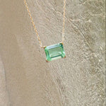 LLUME Jewelry's Seafoam Glass Necklace features a gold chain and a rectangular green gemstone pendant, displayed on textured sand with gentle water ripples, evoking coastal magic.