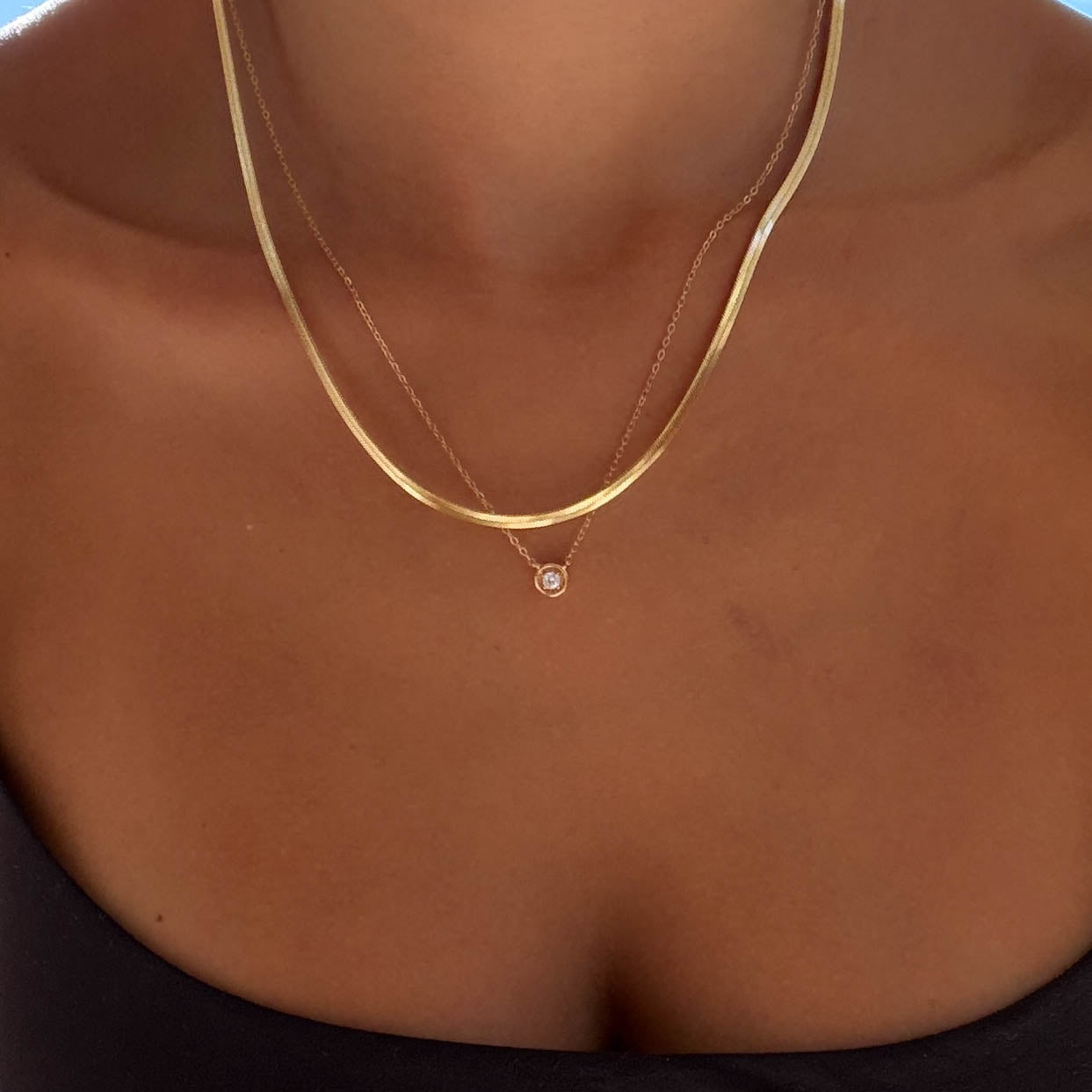 A close-up of a person’s neck and upper chest showcasing the Alexis Set by LLUME Jewelry, layered with a simple chain, against a blue sky. The person is also wearing a black strapless top for an effortless look.