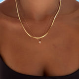 A close-up of a person’s neck and upper chest showcasing the Alexis Set by LLUME Jewelry, layered with a simple chain, against a blue sky. The person is also wearing a black strapless top for an effortless look.