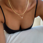 Wearing a black spaghetti strap top and layered gold necklaces from the Alexis Set by LLUME Jewelry, a person leans forward on a white cushion as sunlight streams through a window in the background.