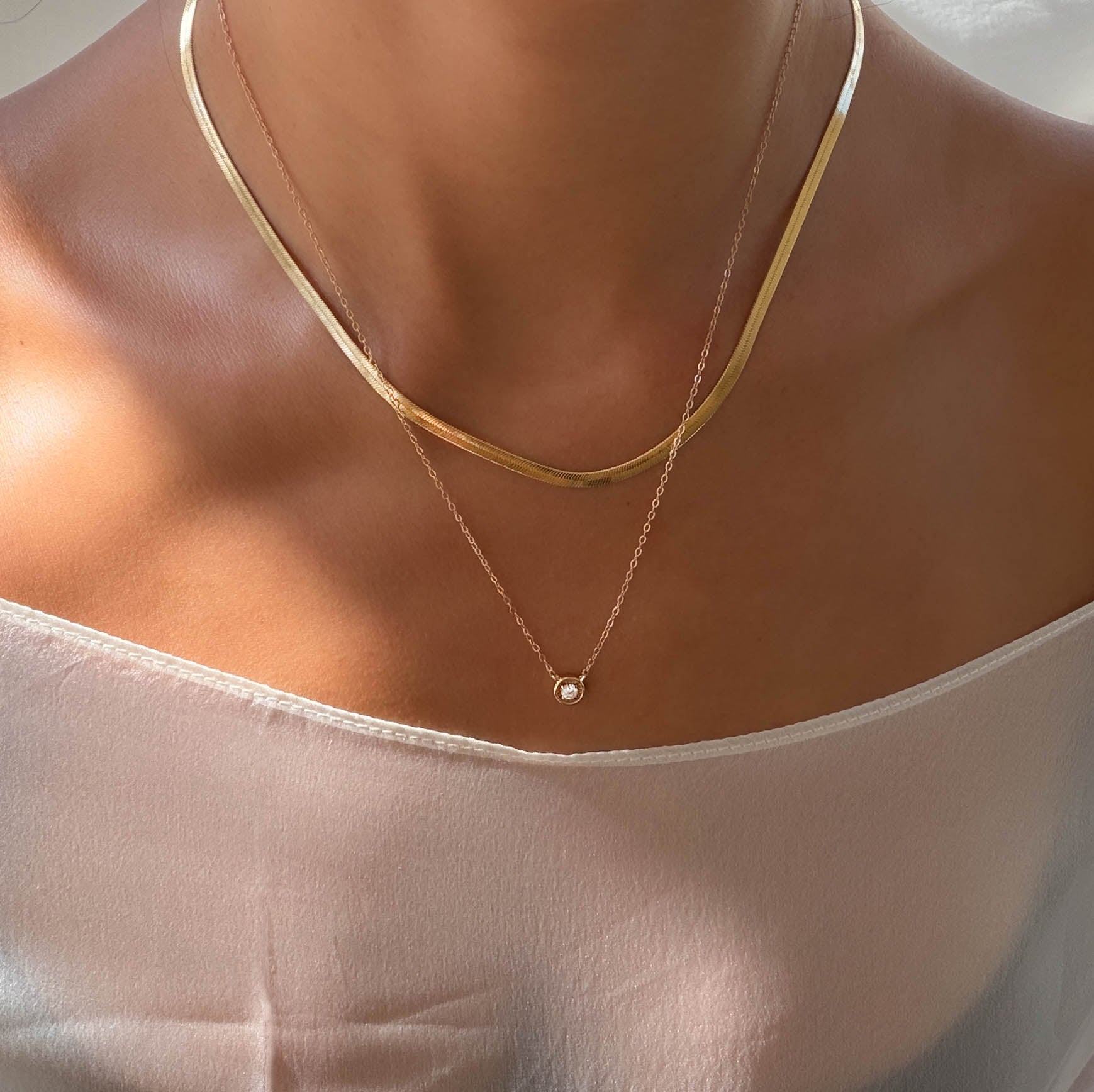 Close-up of a person in a white off-the-shoulder top, wearing the Alexis Set by LLUME Jewelry—a thin gold flat-chain necklace layered with a delicate chain featuring a small round pendant. Soft natural light highlights their skin.