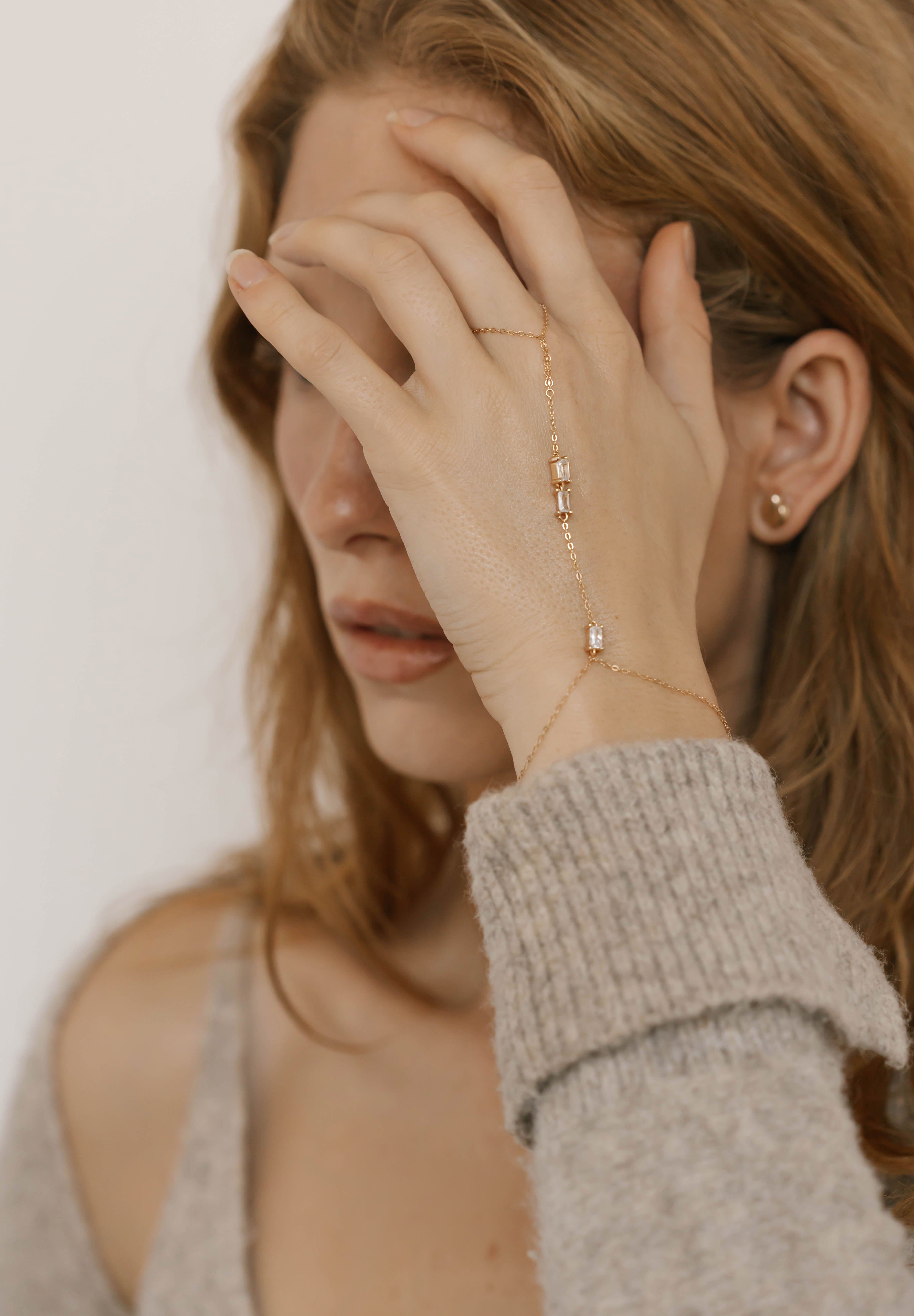 A woman with wavy light brown hair wearing a grey knit sweater covers part of her face with her hand. She wears a delicate gold hand chain bracelet adorned with small gemstones.