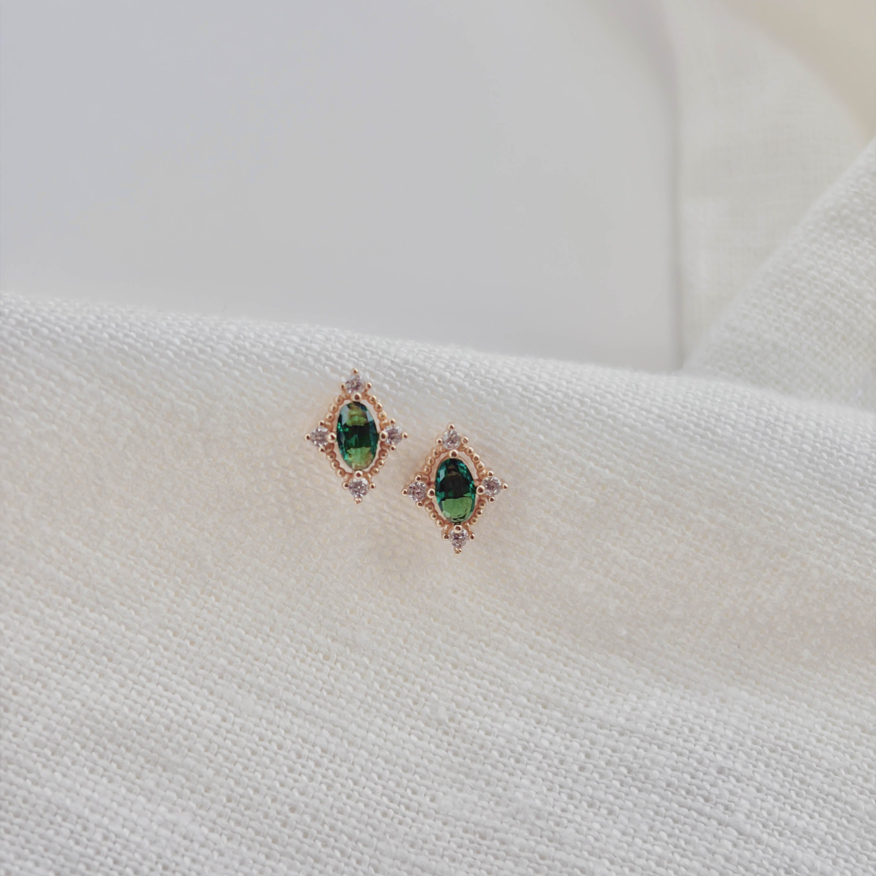 Destiny Earrings by LLUME Jewelry feature gold settings with emerald green studs and clear stones, displayed on a textured white fabric background.