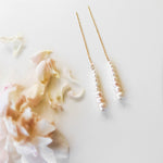 The Elodie Pearl Earrings by LLUME Jewelry feature a line of small white pearls on delicate threaders, elegantly displayed on a white surface beside pale pink flower petals—perfect as wedding jewelry.