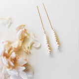 The Elodie Pearl Earrings by LLUME Jewelry feature a line of small white pearls on delicate threaders, elegantly displayed on a white surface beside pale pink flower petals—perfect as wedding jewelry.
