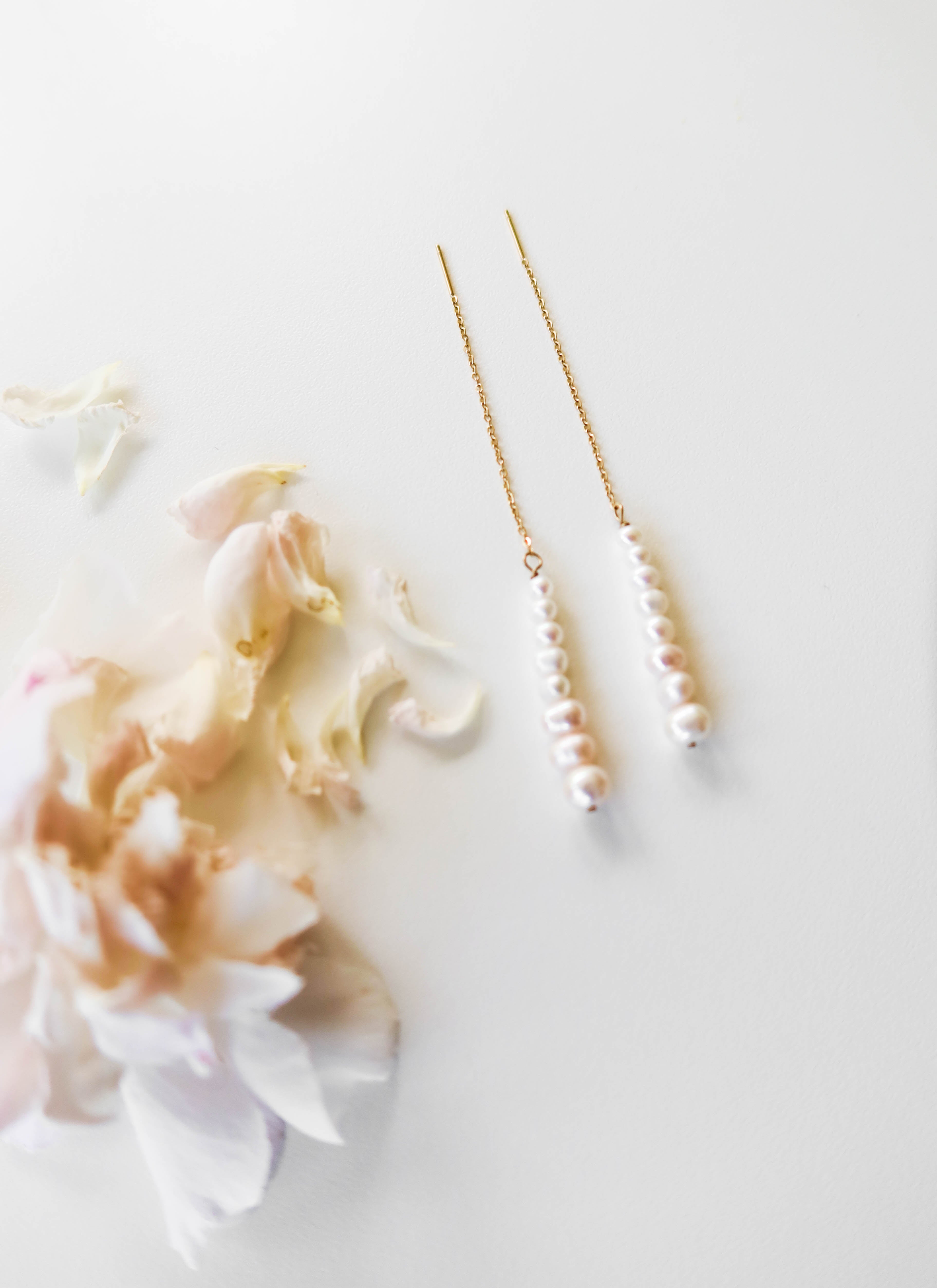 The Elodie Pearl Earrings by LLUME Jewelry feature a line of small white pearls on delicate threaders, elegantly displayed on a white surface beside pale pink flower petals—perfect as wedding jewelry.