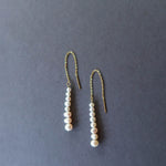 The Elodie Pearl Earrings by LLUME Jewelry feature gold threaders with dangling rows of white pearls, elegantly displayed on a dark grey background—perfect bridal earrings for wedding jewelry.