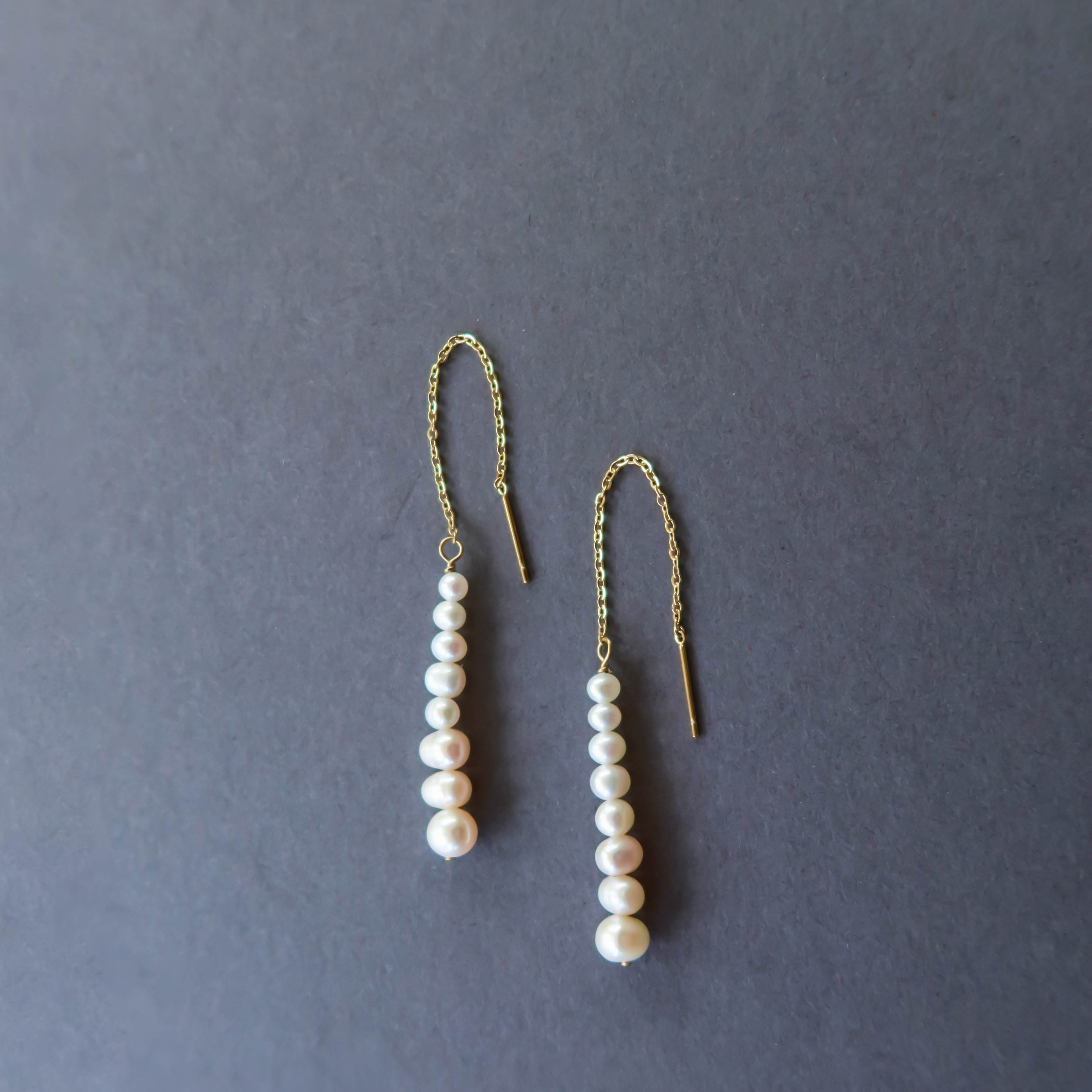 The Elodie Pearl Earrings by LLUME Jewelry feature gold threaders with dangling rows of white pearls, elegantly displayed on a dark grey background—perfect bridal earrings for wedding jewelry.