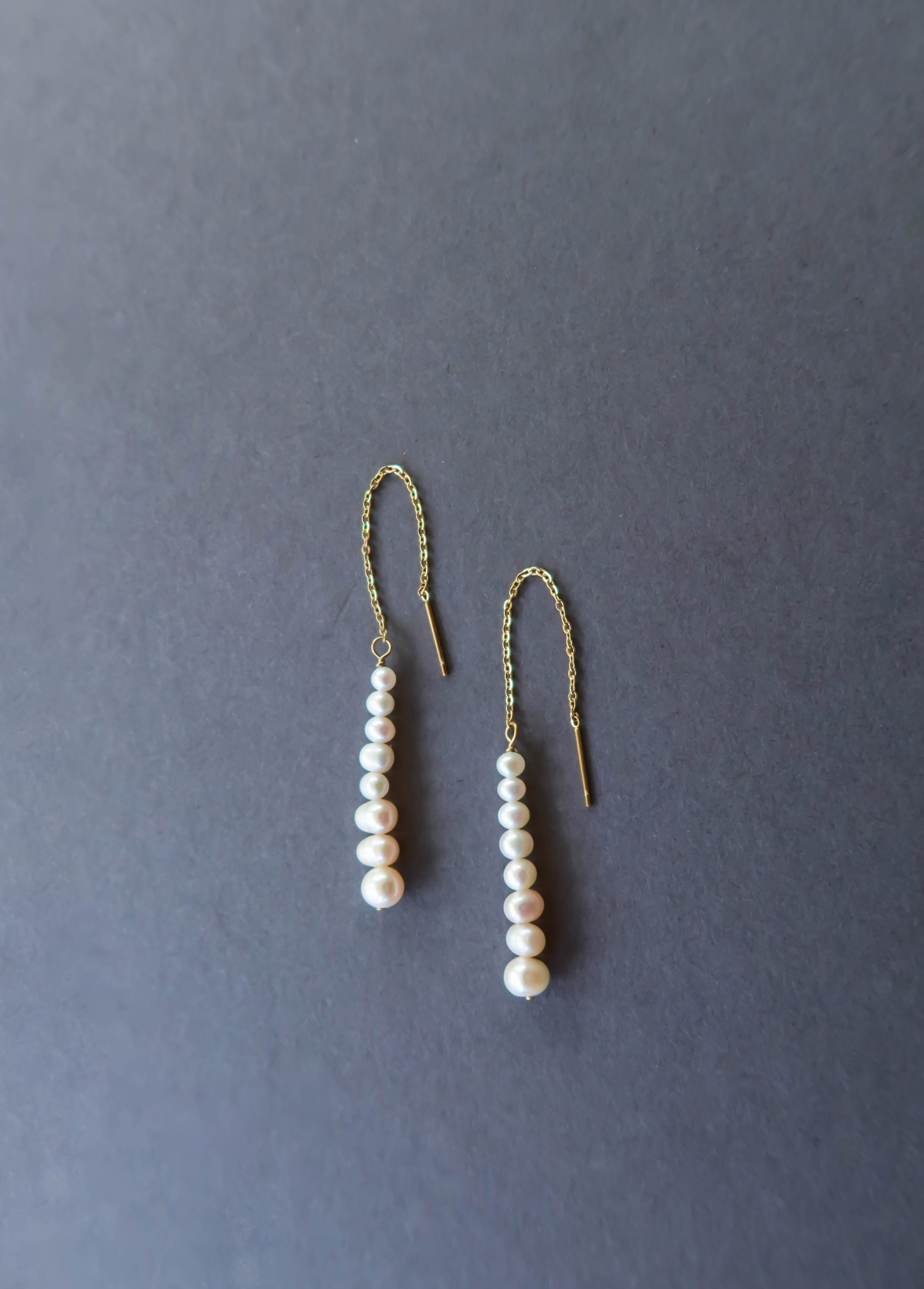 The Elodie Pearl Earrings by LLUME Jewelry feature gold threaders with dangling rows of white pearls, elegantly displayed on a dark grey background—perfect bridal earrings for wedding jewelry.