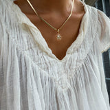 Someone is standing near a window in natural light, wearing a textured white blouse and the LLUME Jewelry Every Occasion Set—a gold snake chain necklace with a sun-shaped pendant.