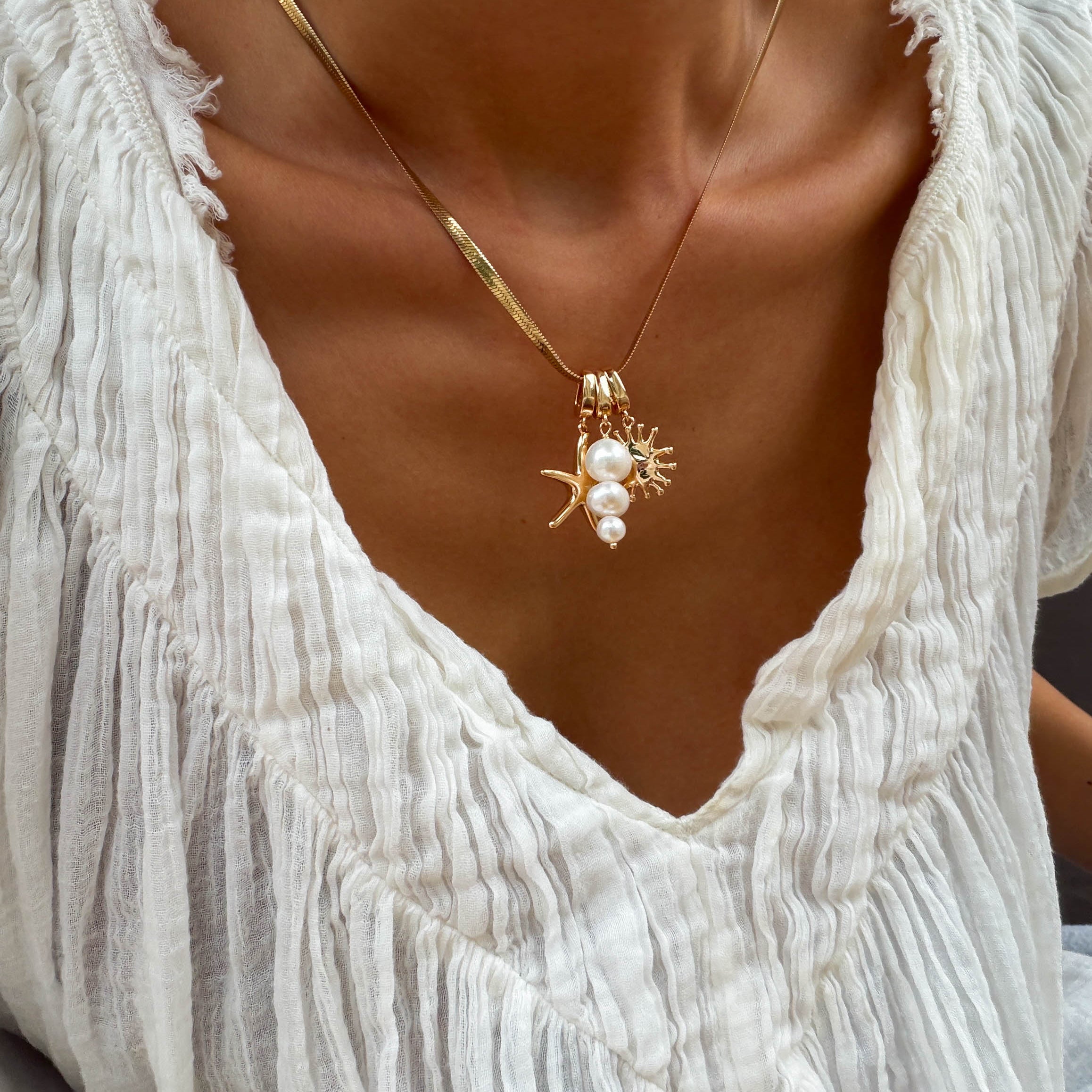 A person wearing a textured white blouse and LLUME Jewelry's Every Occasion Set—a gold snake chain charm necklace with pearl and coral-shaped pendants.