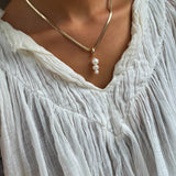 Wearing a crinkled white v-neck blouse, a person accessorizes with the LLUME Jewelry Every Occasion Set—a gold snake chain necklace adorned with four dangling white pearls.