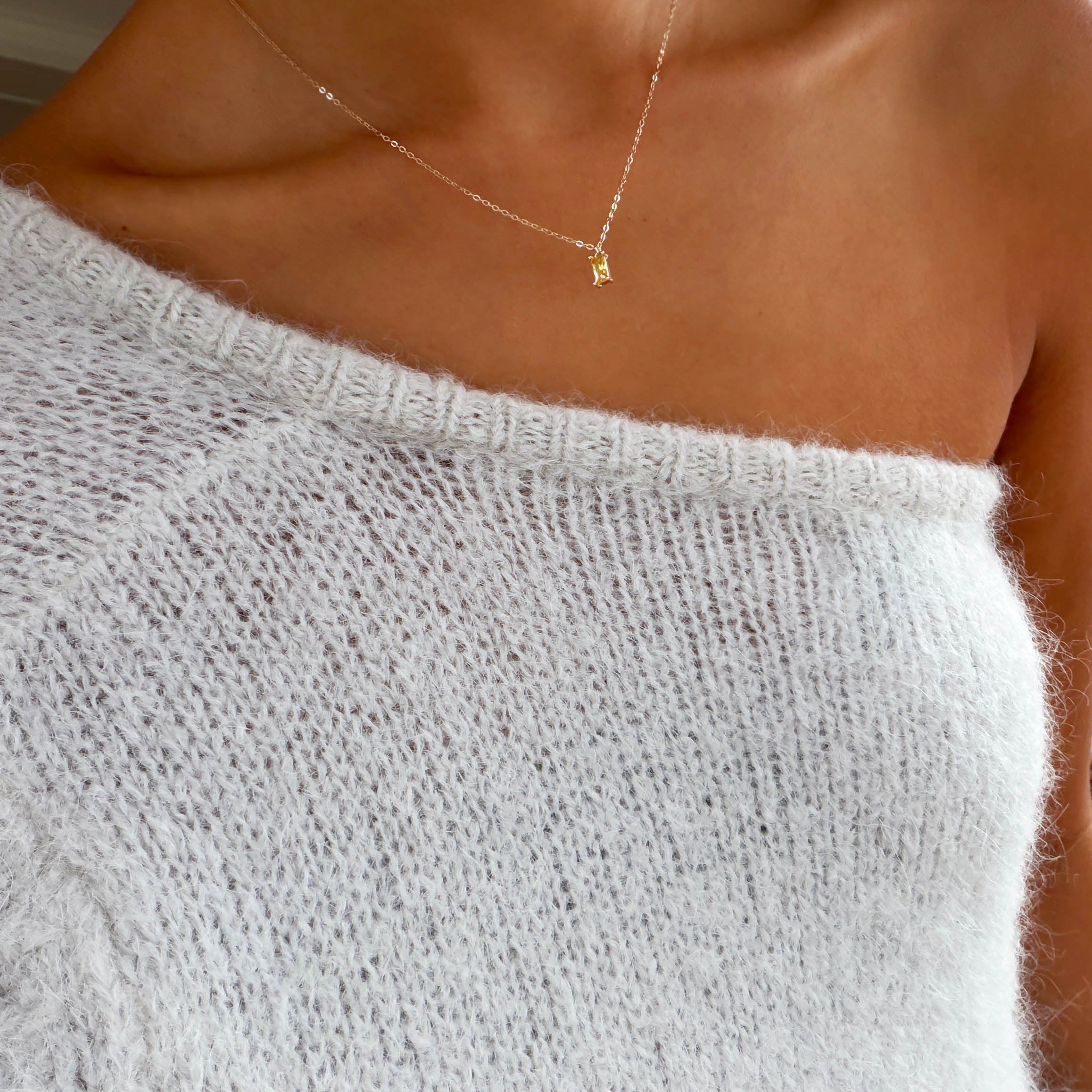 A person wearing a soft white off-the-shoulder sweater and the LLUME Jewelry Lila Necklace, a delicate gold-plated piece with a small pendant, showing bare shoulders and collarbone.