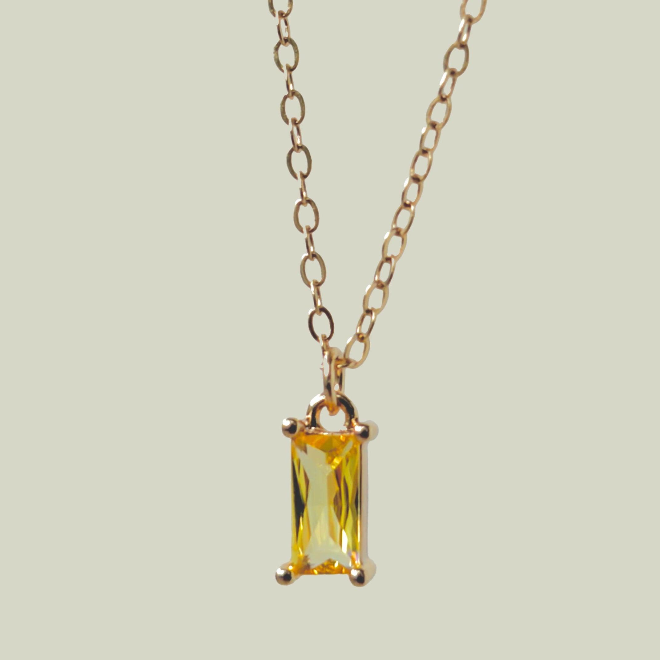 The Lila Necklace by LLUME Jewelry features a gold-plated chain and a rectangular yellow gemstone pendant in a minimalist gold frame, shown on a light beige background.